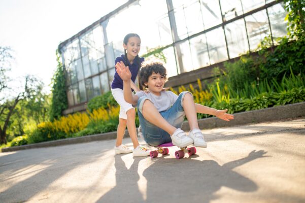 cheerful-cute-dark-haired-girl-helping-friend-ride-skateboard-seated-position-min