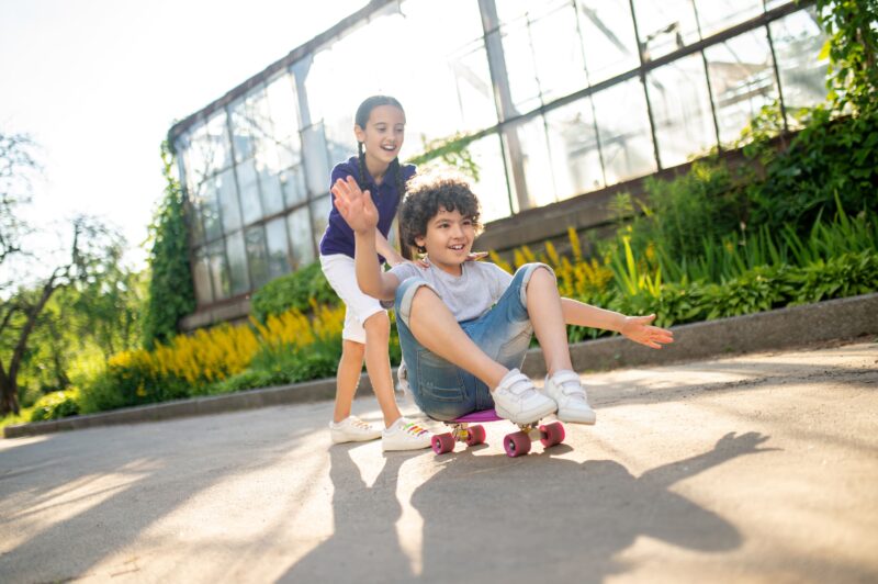 cheerful-cute-dark-haired-girl-helping-friend-ride-skateboard-seated-position-min